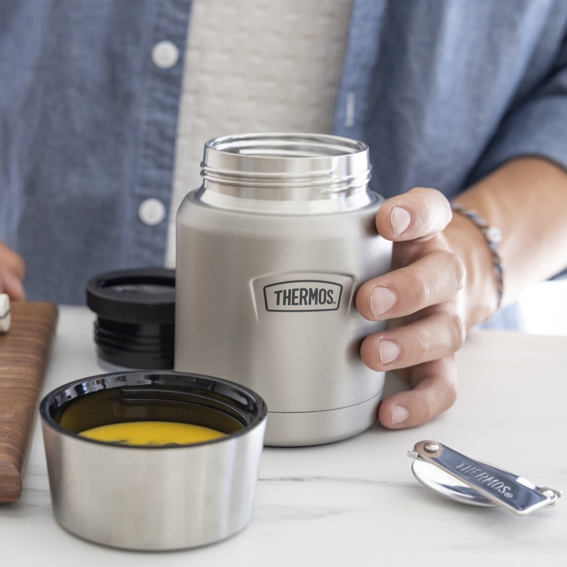 Person holding a Thermos food jar with a lid being used as a bowl on a table