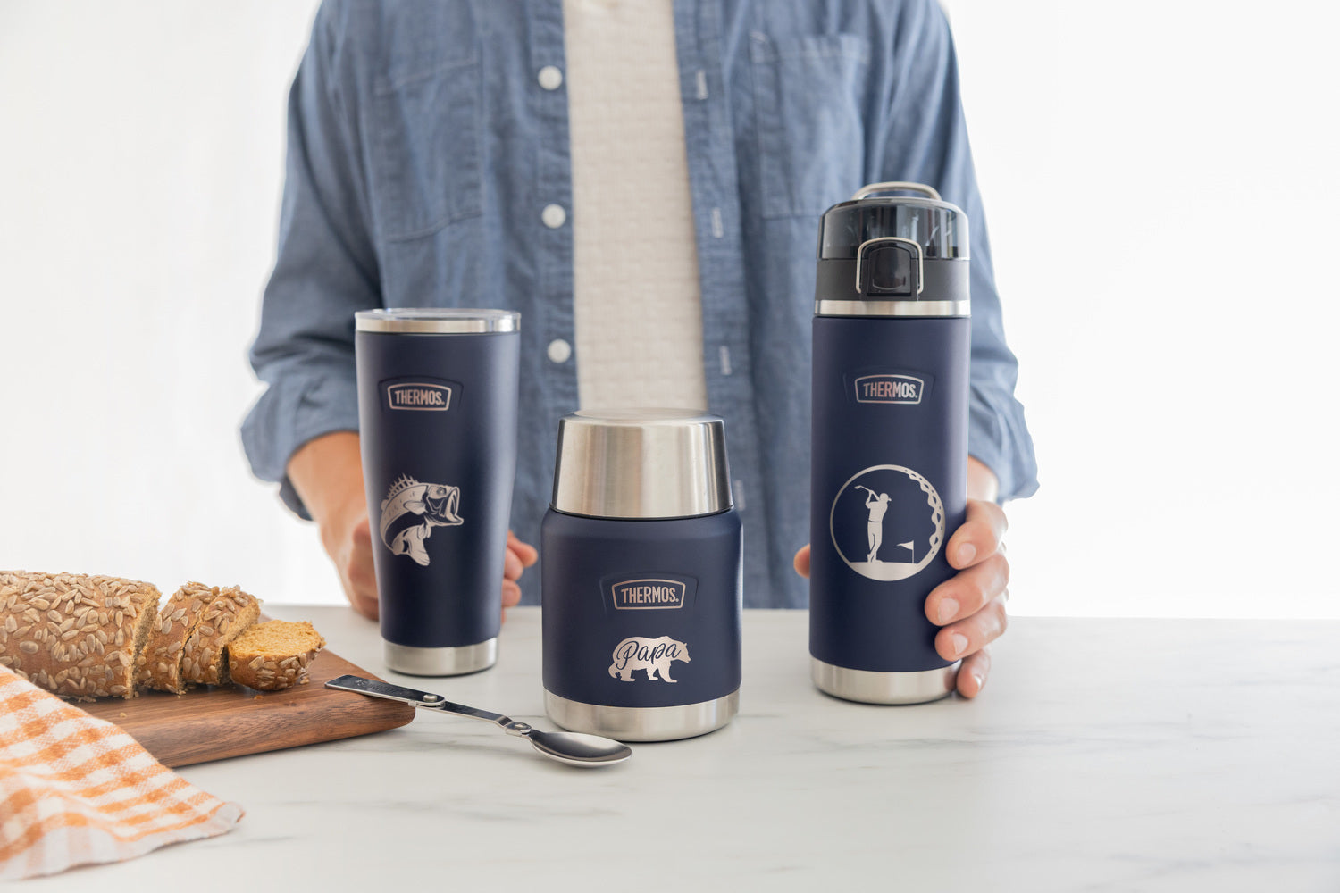 Three Navy Thermos products on a kitchen counter. A tumbler engraved with a bass, a food jar engraved with a "papa" bear, and a water bottle engraved with a person golfing.