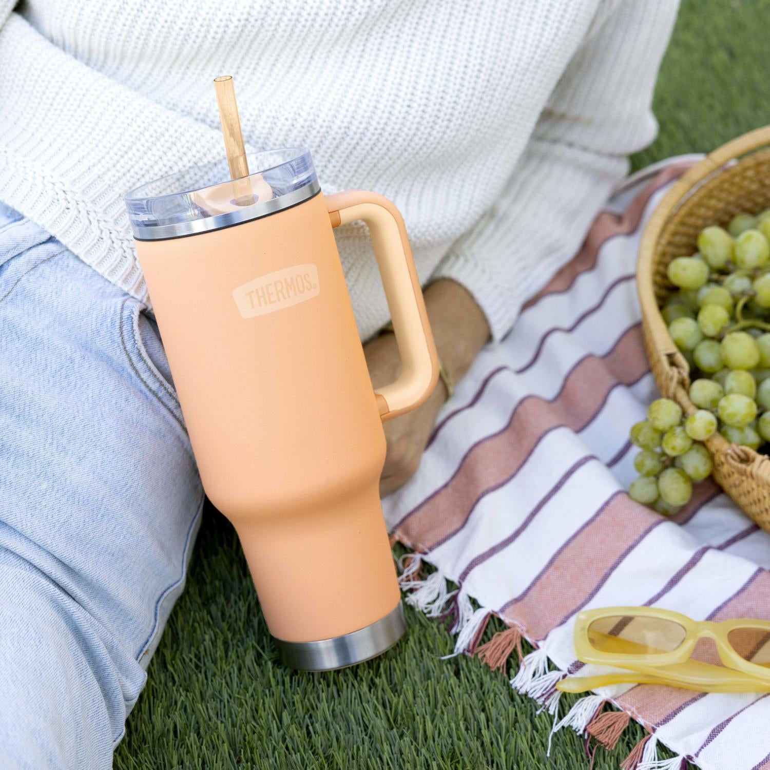 Person holding a peach-colored tumbler with a straw on a picnic blanket with grapes and sunglasses.